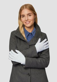 Lichtgrijze gebreide handschoenen met een gestructureerd patroon, gelegd op een persoon die een donkergrijze jas en een blauwe shirt draagt, met gekruiste armen.