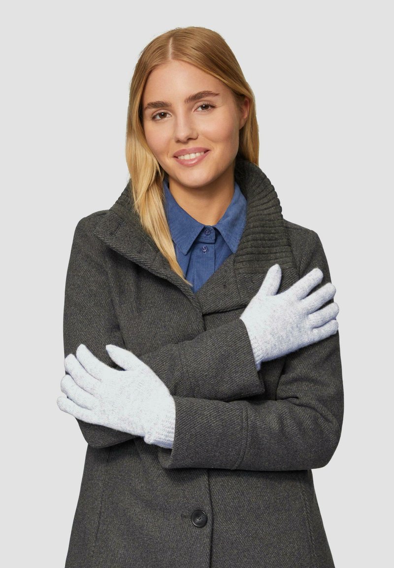 Lichtgrijze gebreide handschoenen met een gestructureerd patroon, gelegd op een persoon die een donkergrijze jas en een blauwe shirt draagt, met gekruiste armen.