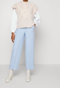 Beżowy sweter dzianinowy z falbaniastymi rękawami i kwiatowym haftem, zestawiony z jasnoniebieskimi spodniami w szerokiej nogawce i kremowymi botkami za kostkę.