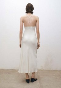 Silke maxikjol i off-white med tunna spagettistilar. Flödande design med en figurnära bodice och en något utfläkt fåll. Svarta klackpumps.