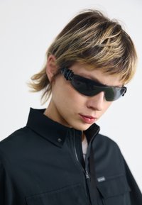 Lunettes de soleil avec des verres teintés foncés, ornées de matériel noir élégant sur les côtés, et d'un design enveloppant et profilé. Le modèle porte une veste noire à col.