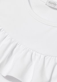 Biała bawełniana koszulka z okrągłym dekoltem i falbanką na dole. Tekstura jest gładka, a szwy widoczne wzdłuż krawędzi.