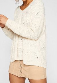 Un pull en laine de couleur crème à coupe ample, avec des motifs ajourés et des poignets côtelés, associé à une jupe beige à boutons sur le devant.