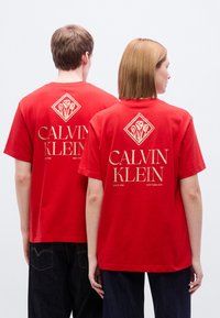 Κόκκινο βαμβακερό μπλουζάκι με χρυσαφένιο λογότυπο στο πίσω μέρος, σχέδιο διαμαντιού και κείμενο "CALVIN KLEIN" με τις φράσεις "ΑΠΟ ΤΟ 1968" και "ΝΕΑ ΥΟΡΚΗ".