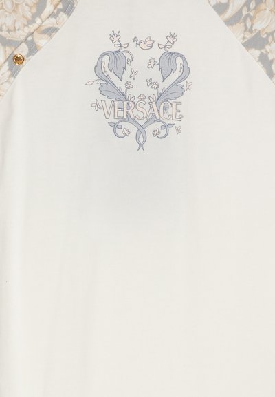 Balta T-kreklu ar gaismas zila printēto dizainu, kurā redzama vārds "VERSACE" ar ziedu akcentiem un teksturētu rakstu piedurknē. Zelta pogu detaļas.