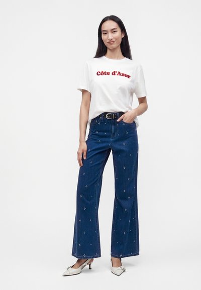 Femme aux longs cheveux foncés portant un t-shirt blanc "Côte d'Azur", un jean bleu taille haute à jambes larges avec de petits motifs, une ceinture noire et des talons blancs.