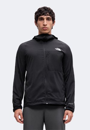 Mand iført en sort hættetrøje med lynlås, med en lynlåslomme på brystet og The North Face-logo på venstre bryst.