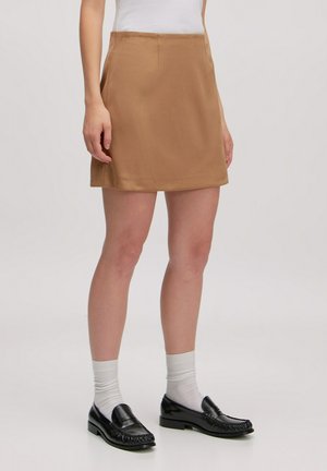 Brauner Minirock aus glattem Stoff, gerader Schnitt und mit einem dezenten Glanz. Kombiniert mit schwarzen Slippern und weißen Socken.