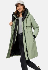 Long green puffer coat with black accents, double-layer hood, zippered pockets, and quilted texture. Model wears a grey shirt and cap.