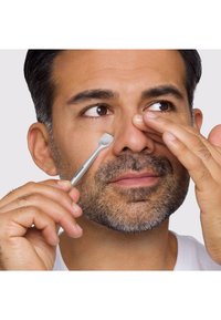Uomo con capelli corti scuri e barba utilizza uno strumento metallico per premere vicino a una narice su sfondo chiaro uniforme.
