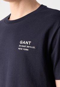 Personne portant un T-shirt bleu marine avec "GANT 32 East 39th St. New York" brodé en blanc sur la poitrine.
