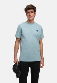 Ljusblå bomulls t-shirt med rund hals och en liten mörk logotyp på vänster bröst. Modellen håller en mörk rutig keps i vänster hand.