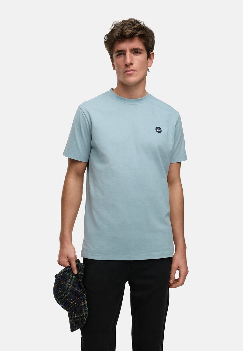 Ljusblå bomulls t-shirt med rund hals och en liten mörk logotyp på vänster bröst. Modellen håller en mörk rutig keps i vänster hand.