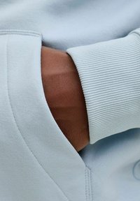 Vaaleansininen pusero resorilla hihansuuissa, jossa ruskea käsi pitää kiinni taskusta. Kangas näyttää pehmeältä ja sileäpintaiselta.