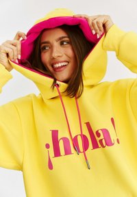 Žltá mikina s ružovým interiérom, s veľkým grafickým nápisom "¡hola!" v ružovej farbe. Sťahovacie šnúrky s kovovými koncami a voľný strih.