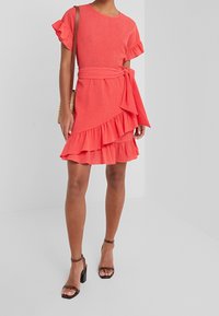 Korálové šaty s volánovými rukávy a lemem, opásané v pase. Vyrobené z texturované tkaniny. Sestavené s hnědými sandály na vysokém podpatku s pásky.