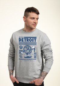 Felpa grigia con un design grafico blu che presenta la scritta "RETRO DETROIT" e un personaggio di basket, con il testo "DUNKING DOWNTOWN" e dettagli in evidenza.