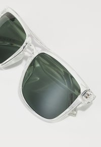 Ochelari de soare din plastic transparent cu lentile verde închis, formă pătrată supradimensionată și accente texturate pe brațe pentru aderență.