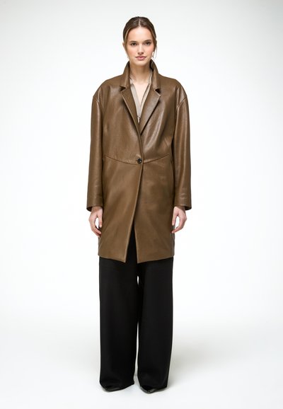 Kobieta w długim brązowym skórzanym płaszczu, na beżowej bluzce i szerokich czarnych spodniach, stojąca na tle jednolitego białego tła.