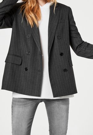 Femme portant un blazer croisé à fines rayures gris foncé sur un pull en maille blanc et un jean gris clair, bras légèrement levés.