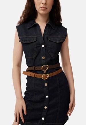 Mujer con vestido sin mangas de mezclilla oscura con botones y dos cinturones, uno marrón y otro beige, ambos con hebillas doradas.