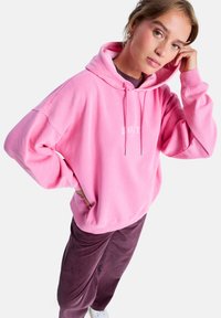 Jeune femme portant un sweat à capuche rose et un pantalon en velours côtelé violet, posant avec une main sur la hanche et l'autre touchant sa tête.