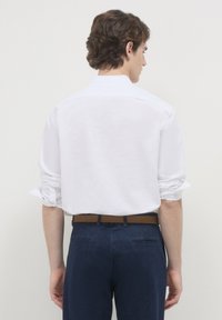 Camicia bianca a maniche lunghe con polsini arrotolati, dotata di un colletto classico. Indossata con pantaloni blu scuro e una cintura marrone. Tessuto liscio.