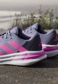 Baskets de sport grises et roses avec une tige texturée, semelle rembourrée et accents roses prononcés. Présente des détails en maille et des rayures emblématiques.