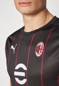 Čierne športové tričko s červenými pruhmi, s bielym logom Puma, kruhovým bielym emblémom a klubovým odznakom AC Milan v červenej a čiernej farbe.