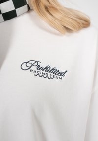 Nærbilde av en hvit genser med "Prohibited Racing Team" brodert i svart skrift nær kragen og et sjakkmønster på skulderen.