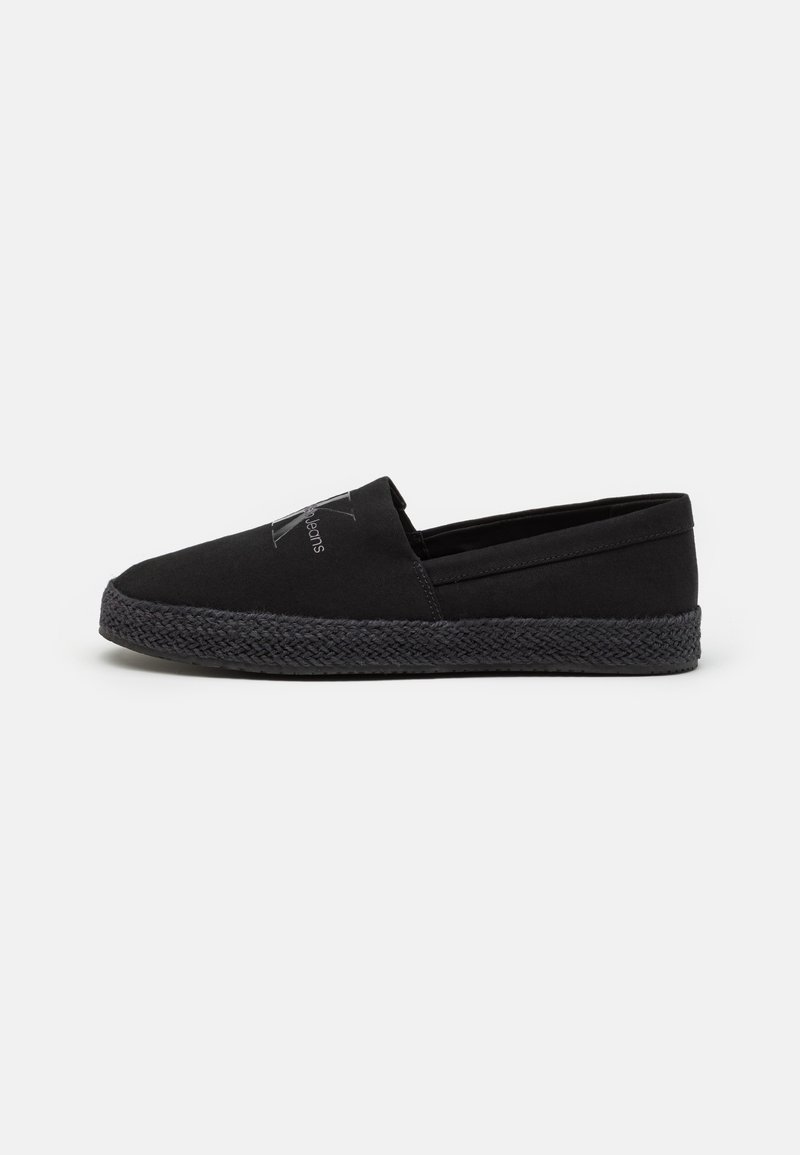 Chaussure décontractée noire à enfiler avec semelle texturée et logo subtil sur la tige, présentée en vue latérale sur fond uni.