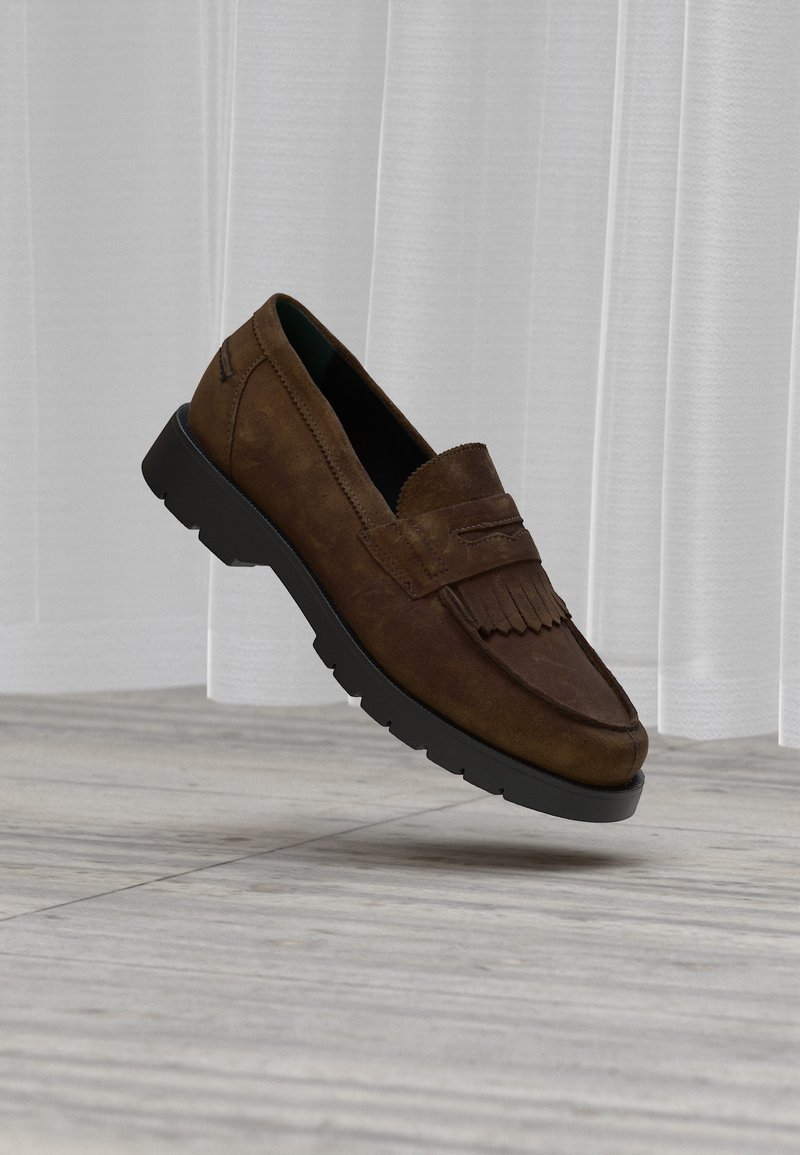 Καφέ δερμάτινο μοκασίνι από σουέτ με διακόσμηση φούντας και μαύρη λαστιχένια σόλα, τοποθετημένο μπροστά σε φως κουρτίνας και φόντο ξύλινης κουζίνας.