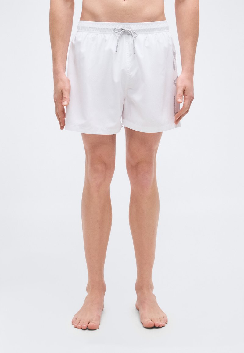 Λευκά μαγιό βερμούδα με κορδόνι στη μέση, μήκους μέχρι τα γόνατα, ελαφρύ υλικό και πλαϊνές τσέπες για ευκολία.