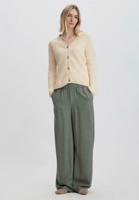 Beige gerippter Cardigan mit Holzknöpfen, kombiniert mit lockeren grünen Hosen und beigen Slippern, vor einem neutralen Hintergrund.