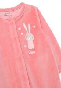 Mono de felpa rosa con un gráfico de conejo, texto "Cutie" y corazones, botones a presión y un acabado suave y texturizado.