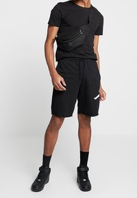 Mustat t-paita ja shortsit pehmeästä kankaasta, käytettyinä mustan vyölaukun kanssa. Mustat lenkkarit, joissa korostetut pohjat. Kaikki esineet minimalistisessa tyylissä.