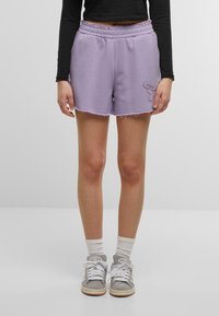 Pantaloni corti tinti di viola chiaro con vita elasticizzata, orlo sfrangiato e dettagli ricamati a forma di farfalla sul lato sinistro. Abbinati a scarpe da ginnastica grigie.