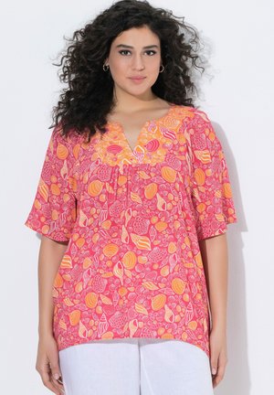 Femme aux cheveux bouclés et foncés portant un chemisier rose et orange à motif coquillages et un pantalon blanc sur un fond blanc uni.