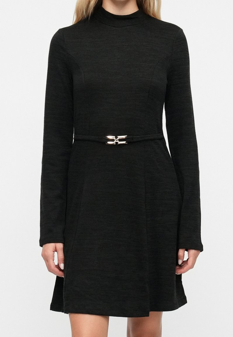 Femme portant une robe noire à manches longues avec un col montant et une fine ceinture à boucle métallique à la taille.