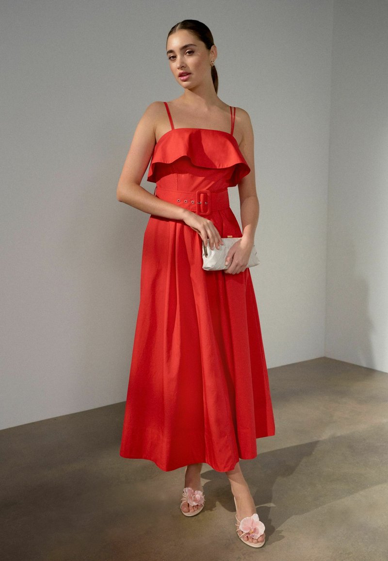Femme debout en robe midi rouge sans manches avec ceinture, tenant une pochette blanche, portant des sandales florales roses, devant un mur clair uni.