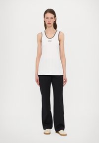 White tank top with black trim and "MM" logo on the chest, paired with black wide-leg trousers and beige sneakers. Minimalist design.
