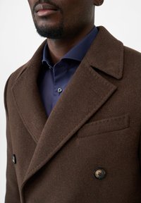 Cappotto di lana marrone con design doppiopetto, grandi rever e una tasca sul petto. Indossato sopra una camicia button-up blu navy.