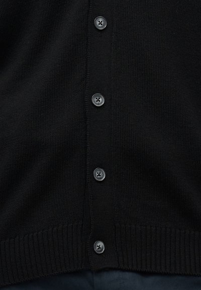 Κοντινό πλάνο μαύρης πλεκτής ζακέτας με τέσσερα σκουρόχρωμα κουμπιά στο κέντρο και ριμπ τελείωμα πάνω από σκούρα παντελόνια.