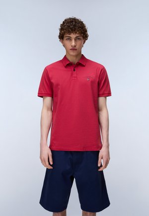 Jeune homme aux cheveux bouclés portant un polo rouge et un short bleu marine, debout sur un fond clair uni.