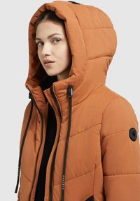 Orange puffjacka med huva, som har svarta snoddar och en svart logopatch på ärmen. Kviltad textur över hela jackan.