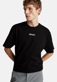 Camiseta de algodón negra de manga corta y cuello redondo. Presenta la palabra "WHAT" en texto blanco en el frente.