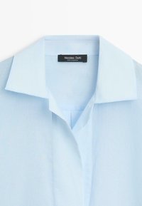 Light blue collared shirt with a V-neck and brand label "Massimo Dutti" sewn inside the neckline.