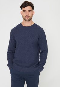 Granatowy sweter o fakturze z wzorem w diamenty, okrągłym dekoltem i ściągaczami przy mankietach. Materiał wydaje się miękki, a fason jest dopasowany.