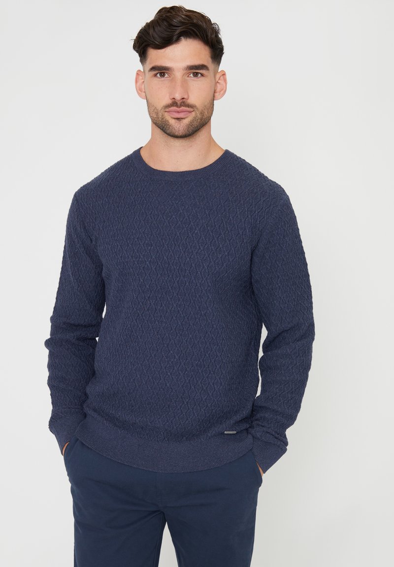 Granatowy sweter o fakturze z wzorem w diamenty, okrągłym dekoltem i ściągaczami przy mankietach. Materiał wydaje się miękki, a fason jest dopasowany.