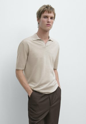 Giovane uomo con polo beige a maniche corte e pantaloni marrone scuro, in piedi con le mani in tasca davanti a uno sfondo chiaro e semplice.